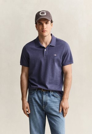 Jeune homme portant une casquette gris foncé avec un G blanc, un polo bleu marine et un jean bleu clair, se tenant devant un arrière-plan uni.