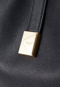 Juodas odinis rankinukas su tekstūruota paviršiumi ir auksinės spalvos trišakiu akcentu, išgraviruotu "kate spade NEW YORK."