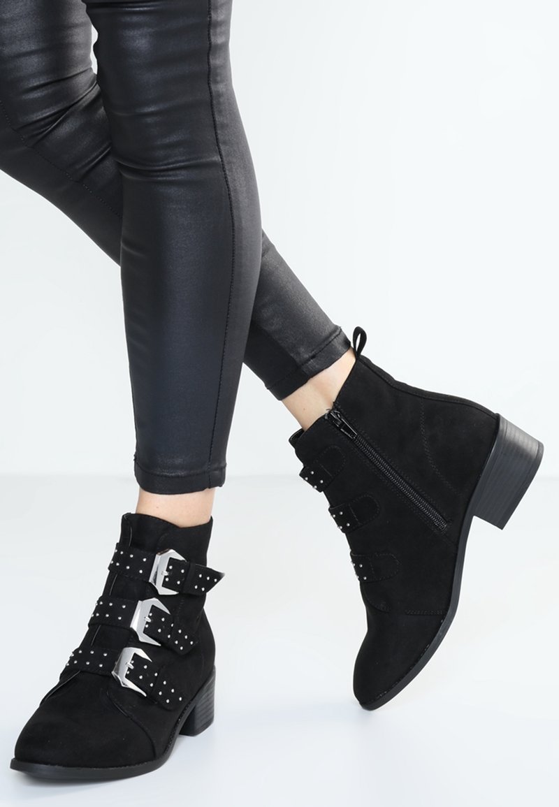 Botas de tobillo de ante negro con tres correas de hebilla con tachuelas y una cremallera lateral, combinadas con pantalones ajustados negros.