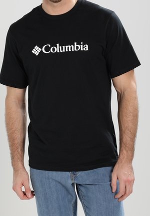 Man die een zwart Columbia T-shirt en blauwe spijkerbroek draagt, staan met ontspannen armen langs het lichaam tegen een egale lichte achtergrond.