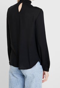 Donna che indossa una blusa nera a maniche lunghe con apertura a chiave sulla schiena e colletto arricciato, abbinata a jeans in denim azzurro chiaro.