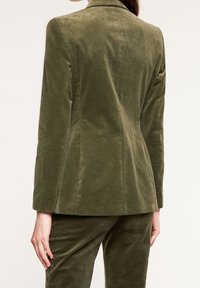 Marynarka ze sztruksu w oliwkowej zieleni o dopasowanym kroju. Posiada rozcięcie z tyłu i gładką teksturę, uzupełnioną eleganckimi, dopasowanymi liniami.