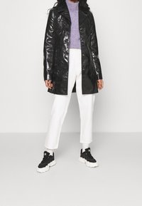 Manteau noir brillant mi-longue avec un col cranté, porté sur un pull violet et un pantalon blanc, assorti à des baskets noires et blanches.