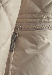 Beige quiltet jakke sektion med en sidelukning og mærke med "ILSE JACOBSEN." Stoffet ser glat ud med en let glans.