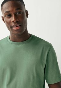 Camiseta de algodón verde con cuello redondo y mangas cortas, que presenta una textura suave y un diseño minimalista. Sin patrones o acentos visibles.