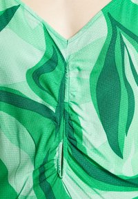 Peça de vestuário de padrão verde com decote em V, apresentando um tecido texturizado, detalhes franzidos e diferentes tons de designs semelhantes a folhas verdes.