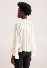 Blusa bianca fluida con maniche a volant e texture plissettata, caratterizzata da un collo alto e una vestibilità comoda, abbinata a jeans scuri.