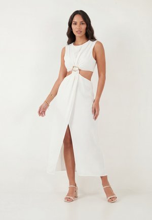 Femme portant une robe midi blanche sans manches avec des découpes à l'avant, un détail anneau doré et une fente frontale, associée à des sandales à talons blocs nude.
