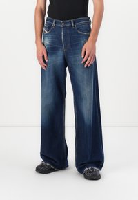 Breechbyxor i blå denim med vida ben och en urblekt finish, fem fickor och synliga sömmar, bar med svarta skor.