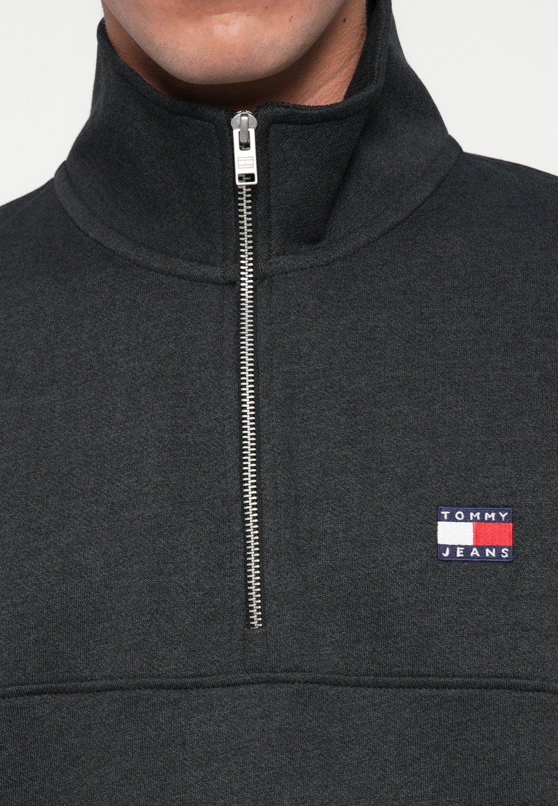 Nahaufnahme eines dunkelgrauen Sweatshirts mit Viertelreißverschluss, silbernem Reißverschluss und kleinem Tommy Jeans Logo-Patch auf der linken Brust.