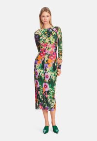 Bunt gemustertes Blumenkleid mit langen Ärmeln, das eine figurbetonte Silhouette und eine Länge bis zur Mitte der Wade aufweist, ergänzt durch grüne spitze Schuhe.
