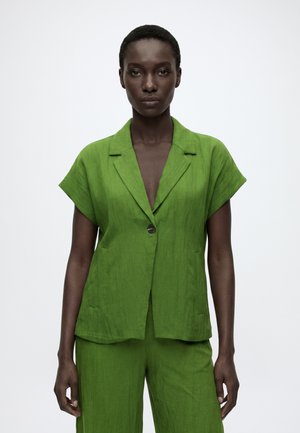 Femme aux cheveux courts portant un blazer vert vif à manches courtes et un pantalon assorti sur un fond blanc uni.