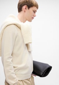 Giovane uomo che indossa un maglione beige e una sciarpa sulle spalle, mentre tiene una grande borsa a busta nera piegata contro uno sfondo bianco.