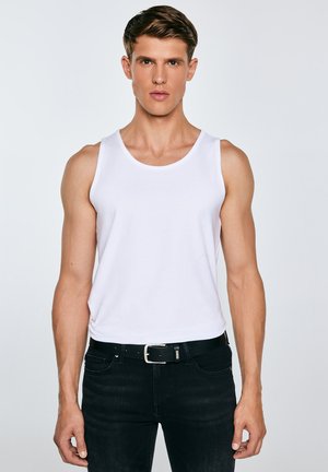 Jauns vīrietis baltā bezroku krekliņā un melnās džinsās ar melnu jostu, stāvot pret vienkāršu gaišu fonu.