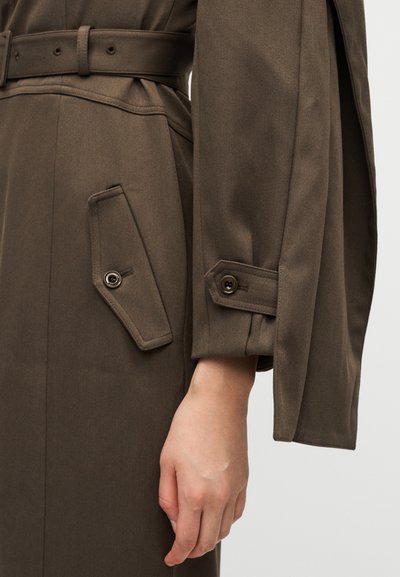 Καφέ καμπαρντίνα με ζώνη, φαρδιές μανίκες, με μεγάλη κεκλιμένη τσέπη και λεπτομέρεια με κουμπί στην μανσέτα, υφασμάτινη υφή.