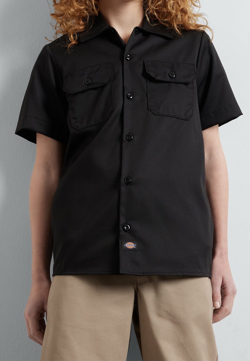 Personne aux cheveux bouclés portant une chemise noire à manches courtes avec des poches sur la poitrine et un pantalon beige, debout devant un mur gris.