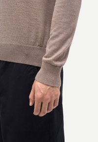 Rechte Hand eines Erwachsenen entspannt neben schwarzer Hose, trägt einen beigefarbenen Pullover mit langen Ärmeln und gerippten Bündchen.