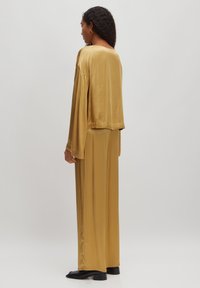 Satin-outfit in senfgelber Farbe mit einem locker sitzenden Oberteil mit weiten Ärmeln und locker fallenden Hosen. Schwarze Schuhe runden den Look ab.