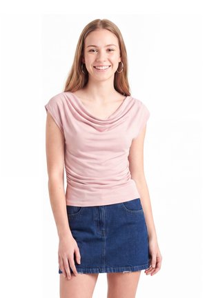 Junge Frau mit glatten, hellbraunen Haaren, die ein rosa drapiertes Oberteil und einen blauen Jeansrock trägt, lächelt vor weißem Hintergrund.