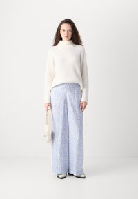 Un pull à col roulé blanc côtelé associé à un pantalon large bleu clair avec un motif ondulé. Tenant un sac à main de couleur claire.