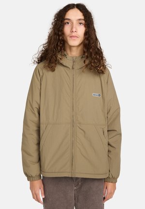 Jongere met lang krullend haar, gekleed in een tan kleurige ritsjack met een klein logo op de borst en elastische manchetten.