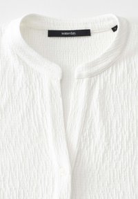 Blusa bianca con texture, dotata di scollo rotondo, chiusura con bottoni e un sottile motivo plissettato. L'etichetta riporta la dicitura "someday."