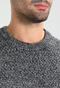 Grau strukturierter Pullover mit rundem Halsausschnitt, der ein gestricktes Muster aufweist. Der Stoff wirkt weich und warm, mit einem dezenten Zweifarbeneffekt.