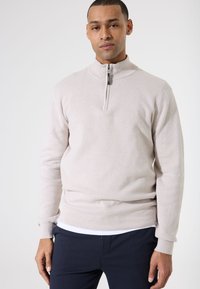 Suéter de media cremallera de color beige claro, hecho de tela texturizada, con un cuello alto y puños y dobladillo acanalados que le dan un aspecto ajustado.