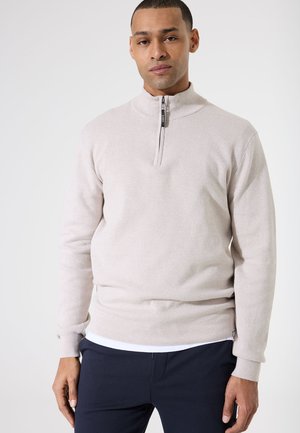 Suéter de media cremallera de color beige claro, hecho de tela texturizada, con un cuello alto y puños y dobladillo acanalados que le dan un aspecto ajustado.