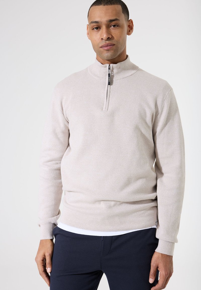 Suéter de media cremallera de color beige claro, hecho de tela texturizada, con un cuello alto y puños y dobladillo acanalados que le dan un aspecto ajustado.