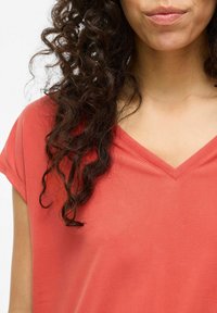 Top de color coral con escote en V y manga corta, fabricado en una tela suave. Presenta un corte holgado y una costura detallada a lo largo del escote.