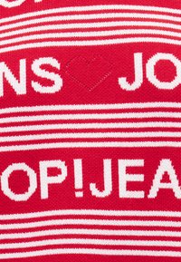 Πλεκτό ύφασμα με κόκκινες και λευκές ρίγες και λευκό κείμενο, περιλαμβάνοντας μια καρδιά ανάμεσα στα γράμματα "NS" και "JO" και μερικές λέξεις "OP! JEANS".