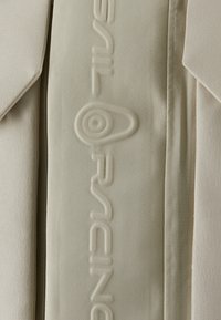 Beige Stoff mit einer strukturierten Oberfläche, der auf einer Seite geprägte "Basil Racing"-Buchstaben aufweist. Klare Linien und einfache Designelemente.