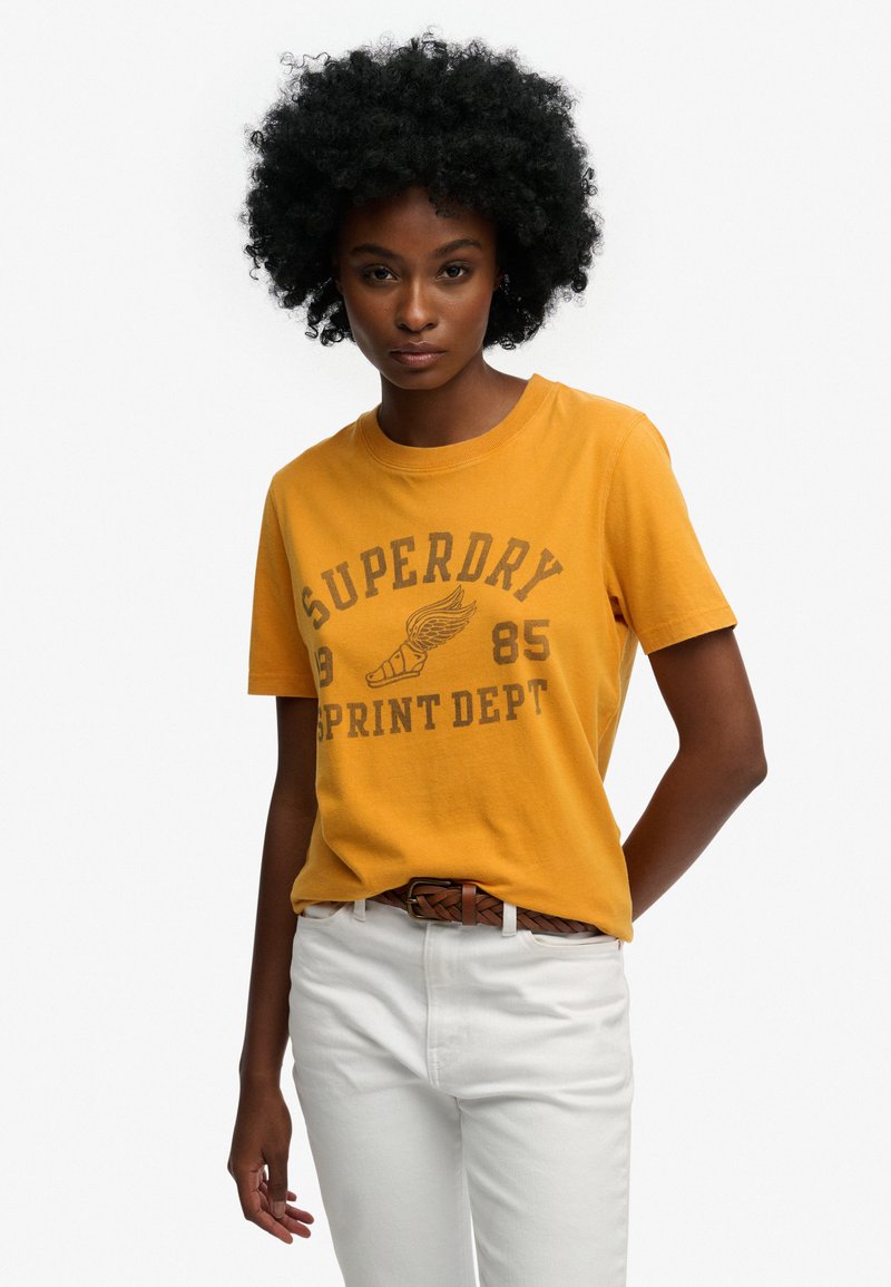 Camiseta de algodón amarilla con mangas cortas y cuello redondo, con un estampado gráfico marrón que incluye el texto "Superdry Sprint Dept". Combinada con pantalones blancos.