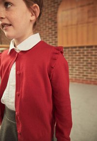 Red cardigan with a ruffled edge, short sleeves, and button closure, worn over a white collared shirt, paired with a gray skirt.