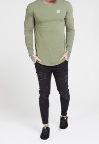 Olivegrünes Langarmshirt mit Rundhalsausschnitt und kleinem weißen Logo; kombiniert mit schwarzen, zerrissenen Skinny-Jeans und schwarzen Sneakers.