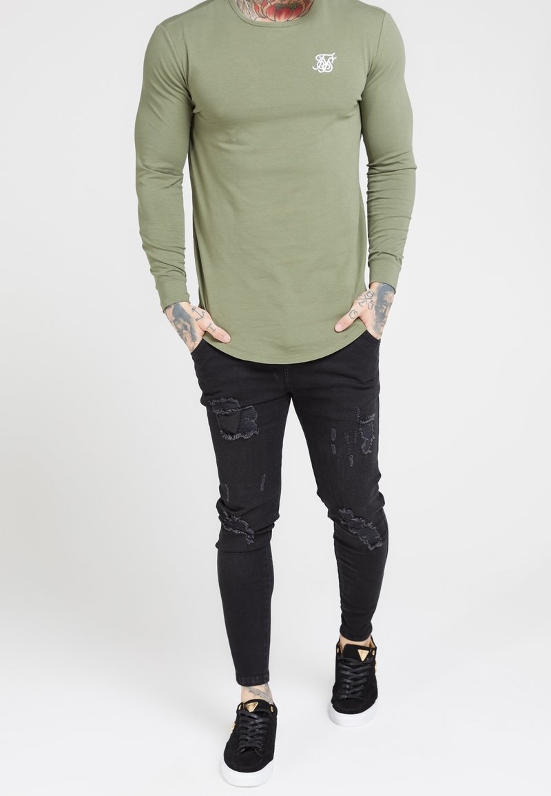 Olivegrünes Langarmshirt mit Rundhalsausschnitt und kleinem weißen Logo; kombiniert mit schwarzen, zerrissenen Skinny-Jeans und schwarzen Sneakers.