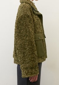 Olivengrøn yderjakke, der kombinerer struktureret, fluffy fleece med glat stof. Har en stor knaplomme og trykknaplukning.