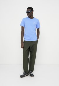 Ανοιχτό μπλε κοντομάνικη μπλούζα Nike με λευκό λογότυπο, συνδυασμένη με ελιάς cargo παντελόνι και μαύρα αθλητικά παπούτσια. Αξεσουάρ περιλαμβάνουν μπλε γυαλιά ηλίου.