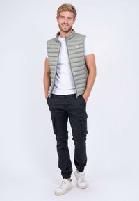 Gilet matelassé gris clair avec coutures verticales, fermeture éclair et logo. Porté par-dessus un t-shirt blanc, associé à un pantalon cargo noir et des baskets blanches.