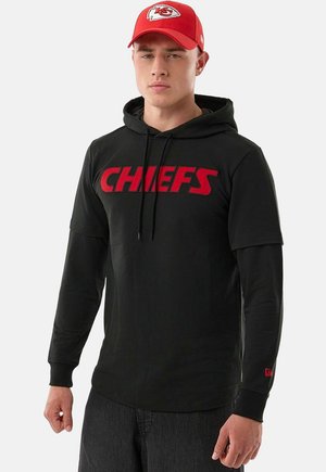Giovane uomo con felpa nera dei Chiefs con scritte rosse e cappellino da baseball rosso dei Kansas City Chiefs, in piedi davanti a uno sfondo semplice.