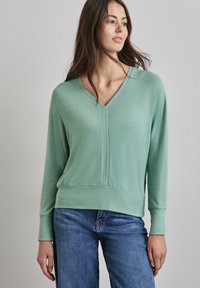 Femme portant un pull à col V à manches longues vert menthe avec des poignets côtelés et un jean bleu, se tenant contre un fond gris clair uni.