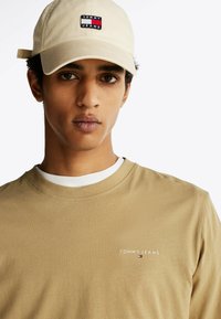 Beżowa koszula z długim rękawem z białym wewnętrznym kołnierzykiem, z małym haftowanym logo "TOMMY JEANS" oraz bawełnianą fakturą. Beżowa czapka z daszkiem z logo.