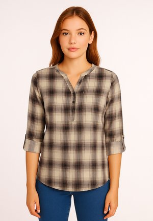 Young woman with straight brown hair wearing a beige and black plaid buttoned shirt and blue jeans standing against a plain background.