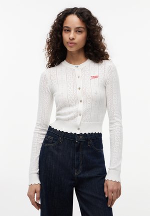 Junge Frau trägt eine weiße, geknöpfte Strickjacke mit dem „tommy jeans“ Logo und dunkle, hoch taillierte Jeans vor einem schlichten Hintergrund.