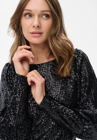 Vrouw met lichtbruin haar die een zwart, glinsterend shirt met lange mouwen draagt, en iets opzij kijkt met haar handen naast haar gezicht.