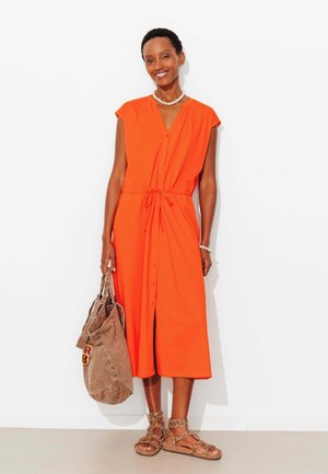 Femme souriante, portant une robe midi orange vif boutonnée avec taille à cordon, sandales imprimé léopard, collier et bracelet de perles, tenant un sac marron.