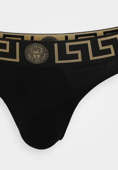 Schwarze Baumwollslips mit einem dekorativen goldenen Bund im griechischen Schlüssel-muster und einem zentralen Medusa-Logo. Glattes Material mit leichtem Glanz.