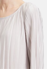 Hellbeige, plissierte Bluse mit einem runden Halsauschnitt, fließenden Ärmeln und glatter Textur. Mit vertikalem Streifenmuster durchzogen.