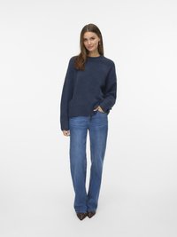 Eine Frau trägt einen dunkelblauen, oversized Pullover, blaue gerade Jeans und spitze, braune, mit Nieten besetzte Schuhe und steht vor einem weißen Hintergrund.